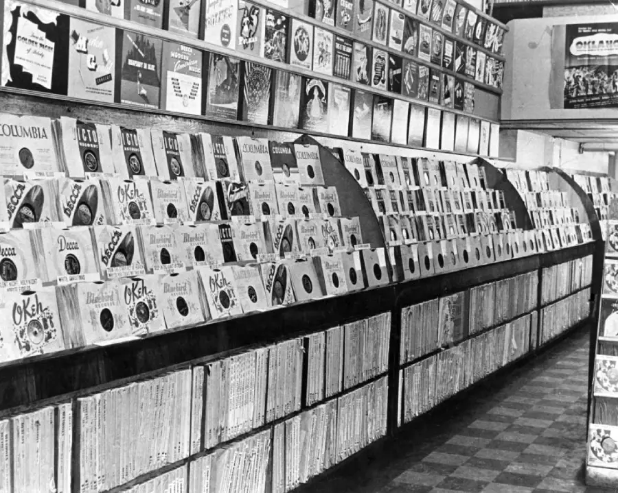 Black-and-white photo of a record store with rows of vinyl records displayed on shelves and album covers visible, creating a nostalgic, vintage atmosphere. The floor has a checkered pattern.