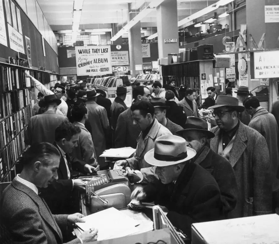 A crowded record store in the 1950s, with people in hats and coats browsing and buying albums at the counter. Shelves are filled with vinyl records, and signs advertising deals hang overhead.