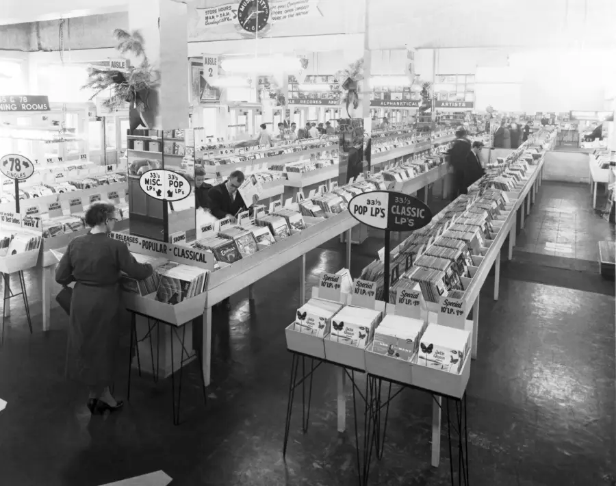 Black and white photo of a large record store with rows of vinyl records, signs for “POP LPS” and “CLASSIC LPS,” and several people browsing the aisles under bright overhead lights.