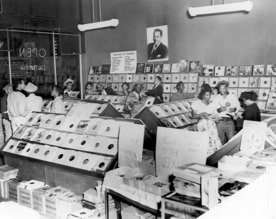 Black-and-white photo of a busy vintage record store with shoppers browsing vinyl records, albums displayed on the walls, a portrait above, and handwritten signs visible among the record bins.