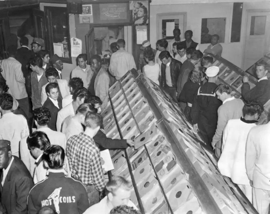 Black and white photo of a crowded record store with people browsing through bins of vinyl records. Most are men, dressed in 1950s-1960s attire, looking through albums.