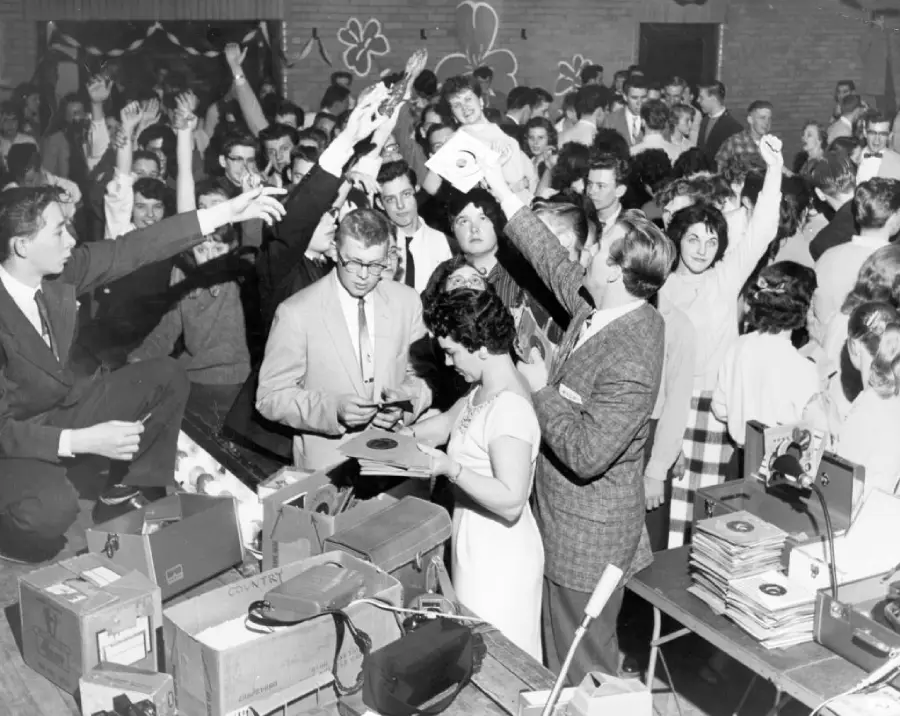 A black-and-white photo shows a crowded room of excited people, many reaching out for vinyl records being handed out by formally dressed individuals at a table covered with records and boxes.