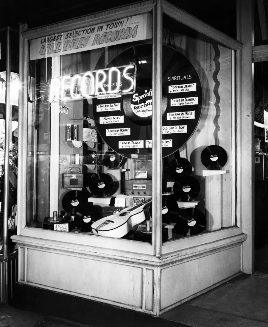 A vintage record store display window shows vinyl records, a guitar, record players, and signs advertising “LARGEST SELECTION IN TOWN” and “SPIRITUALS.” Neon “RECORDS” sign hangs above.