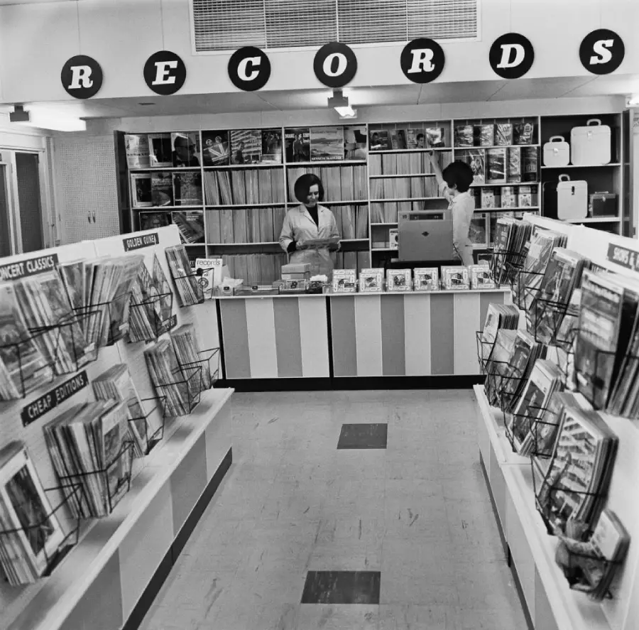 Black-and-white photo of a vintage record store. Two women stand behind the counter, and LPs are displayed in bins and shelves. Large round signs above the counter spell out “RECORDS.”