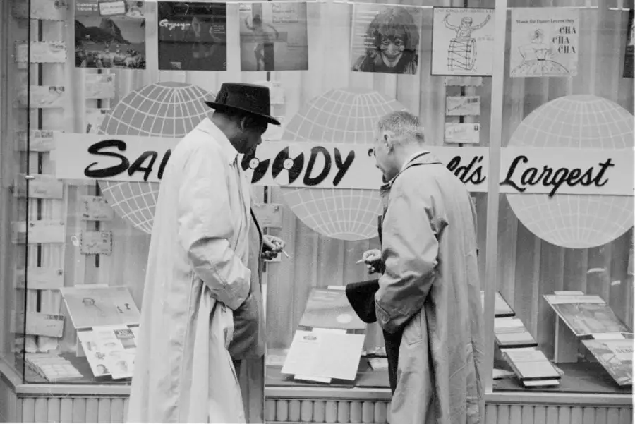 Two men in coats stand in front of a shop window display, looking at documents or papers. The window is decorated with signs reading “Sam Moody World’s Largest” and various posters and flyers.