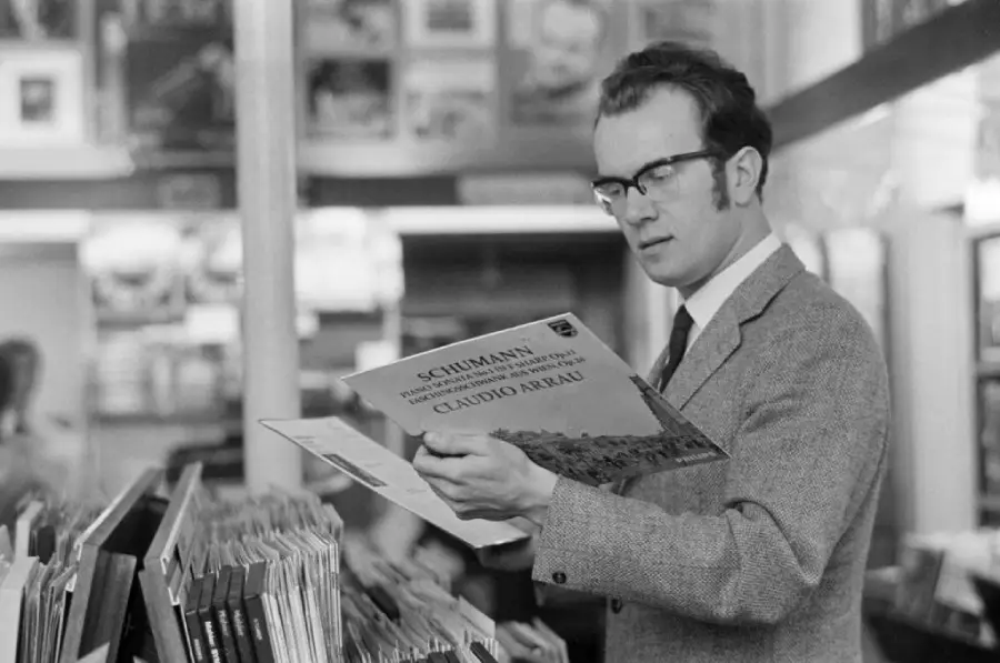 A man wearing glasses and a suit looks at a Schumann record in a record store, surrounded by shelves of vinyl albums.