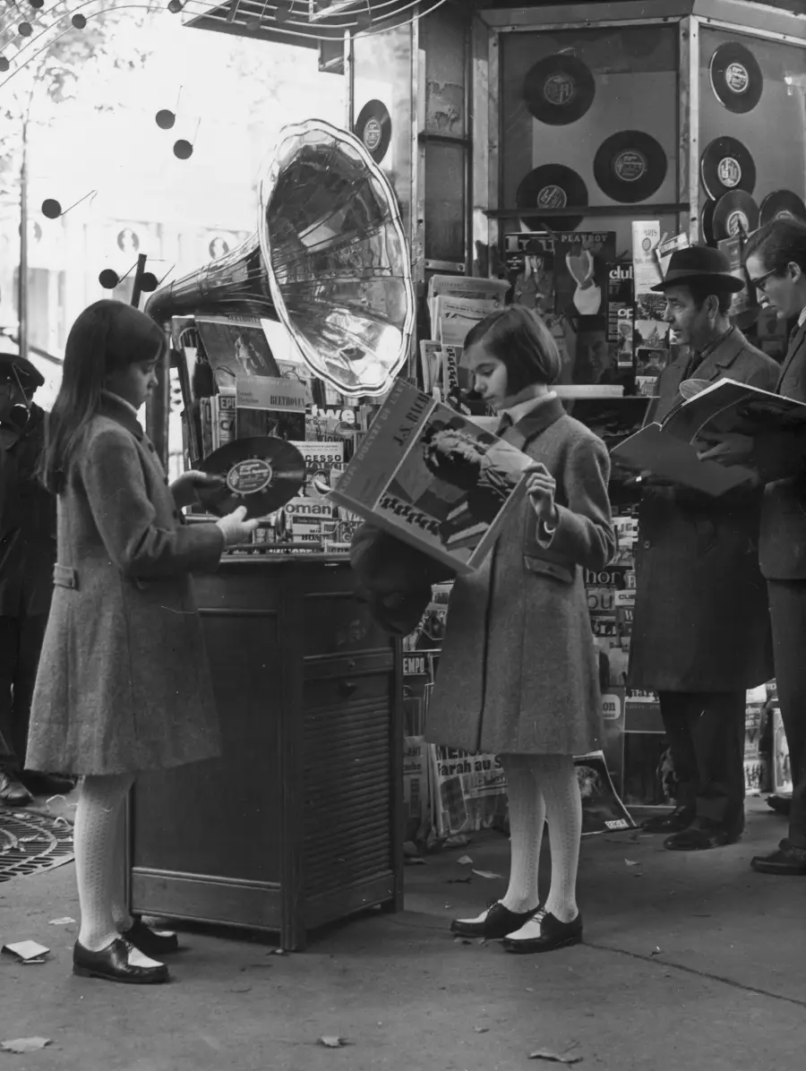 Two girls in coats and tights look at vinyl records near a large gramophone outside a newsstand, while men in hats read newspapers in the background.