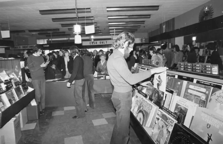 Black and white photo of people browsing records in a busy vintage record store; one person in the foreground flips through albums while others shop and talk in the background.