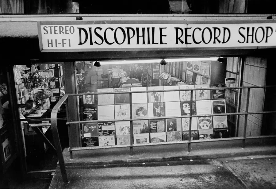 Black-and-white photo of the front of Discophile Record Shop. Vinyl records and posters are displayed in the window, and a sign above the entrance reads “Stereo Hi-Fi Discophile Record Shop.”