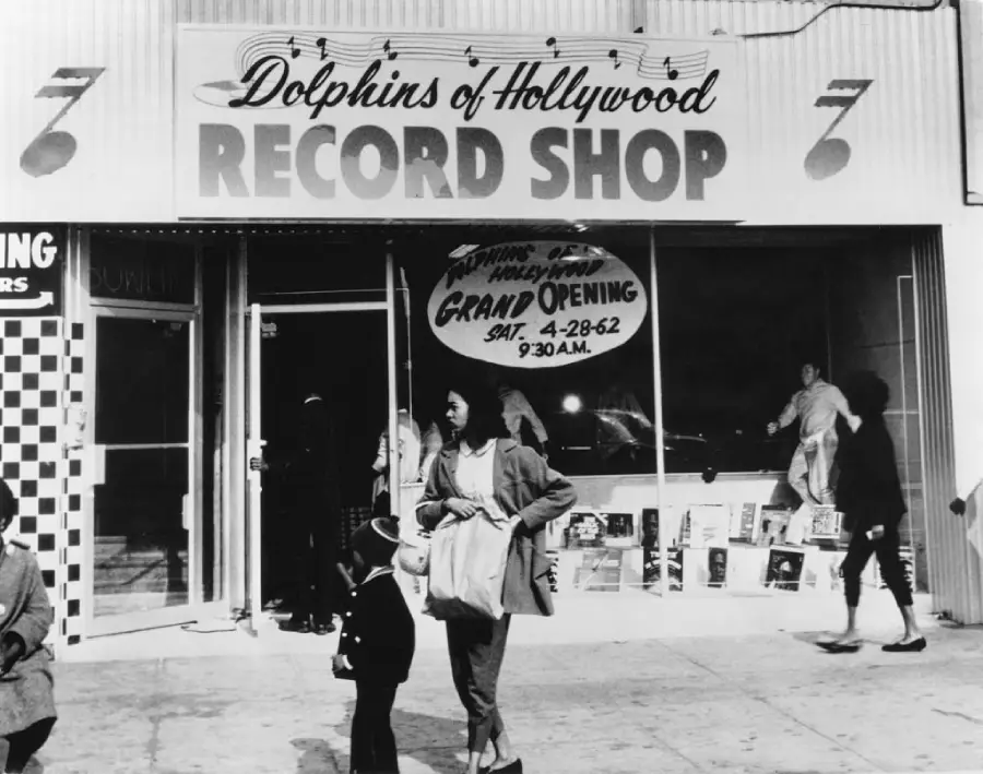 Black and white photo of Dolphins of Hollywood Record Shop with “Grand Opening” sign in the window. Several people stand or walk near the storefront, and records are displayed inside.