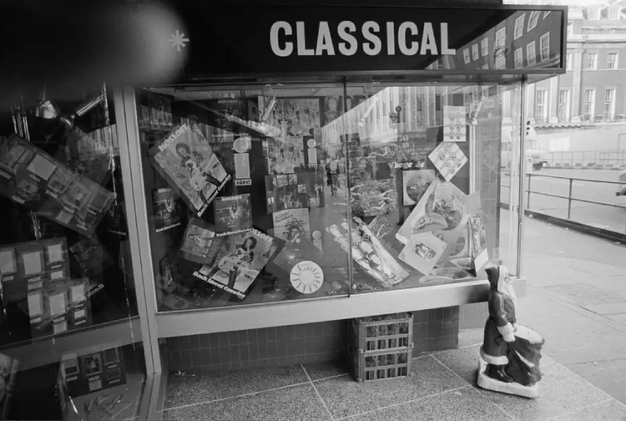 Black and white photo of a shop window labeled “CLASSICAL,” displaying records with illustrated covers. A Santa Claus figure sits on the pavement outside, and a crate is placed near the glass. Part of a finger covers the top left.