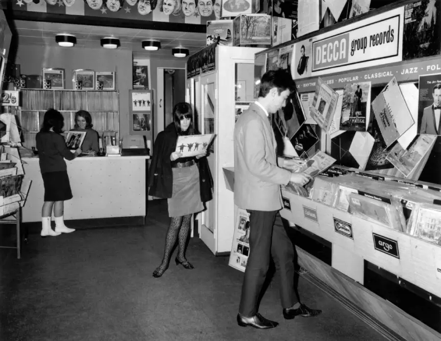 Black-and-white photo of three people browsing vinyl records in a vintage record store with posters on the walls and “DECCA group records” sign above record bins.