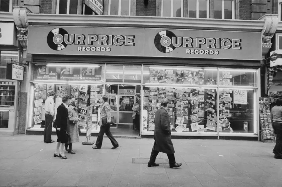 Black and white photo of people walking past a storefront called “Our Price Records,” featuring large display windows filled with posters and records.