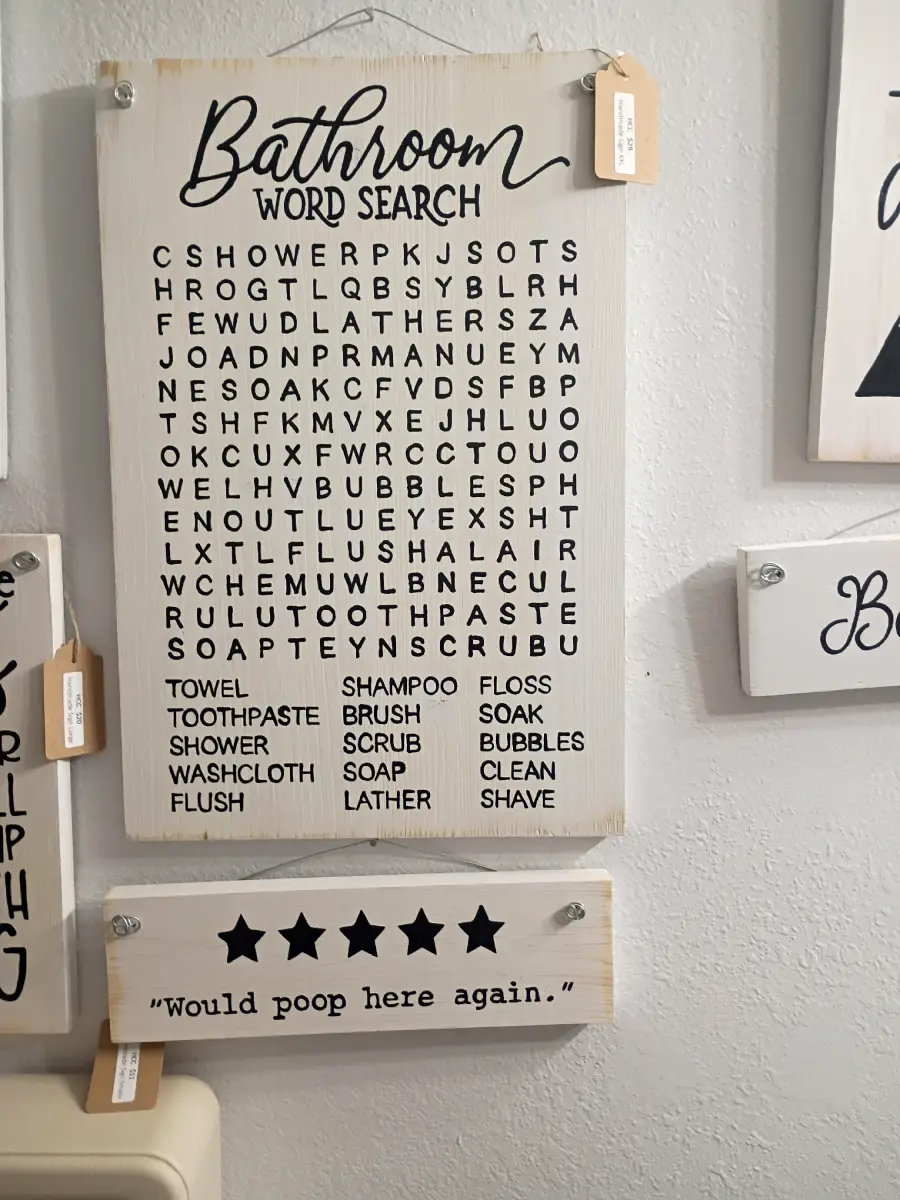 A bathroom-themed word search sign lists items like towel, shampoo, and soap. Below it, a sign with five stars humorously reads, “Would poop here again.” Both signs are mounted on a wall.
