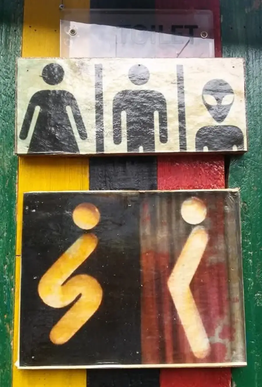 Two bathroom signs: the top shows three abstract figures, the bottom shows stick figures—one squatting (for women), one standing (for men). Both signs are mounted on a colorful striped wall.