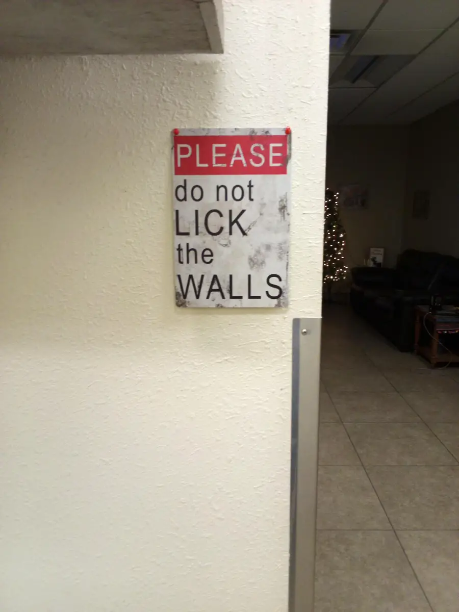 A sign on a wall reads “PLEASE do not LICK the WALLS” in black and red text, with some smudges and marks visible on the sign.