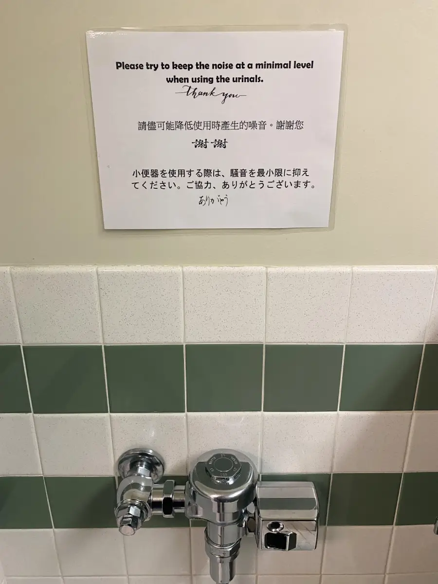 A sign above a urinal asks users to keep noise to a minimum and includes text in English, Chinese, and Japanese. The urinal and tiled wall are visible below the sign.
