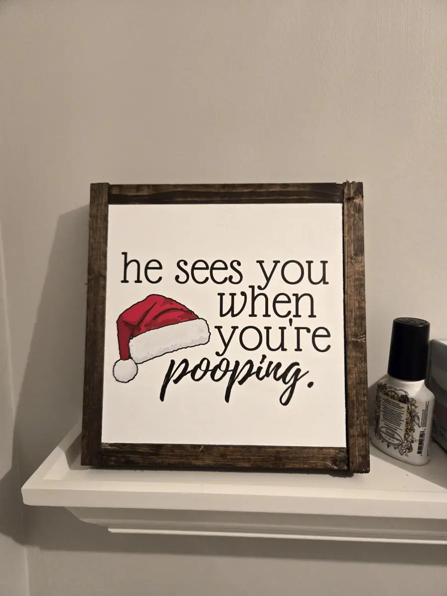 A framed sign with a Santa hat illustration reads, “he sees you when you’re pooping.” The sign sits on a white shelf next to a bottle.