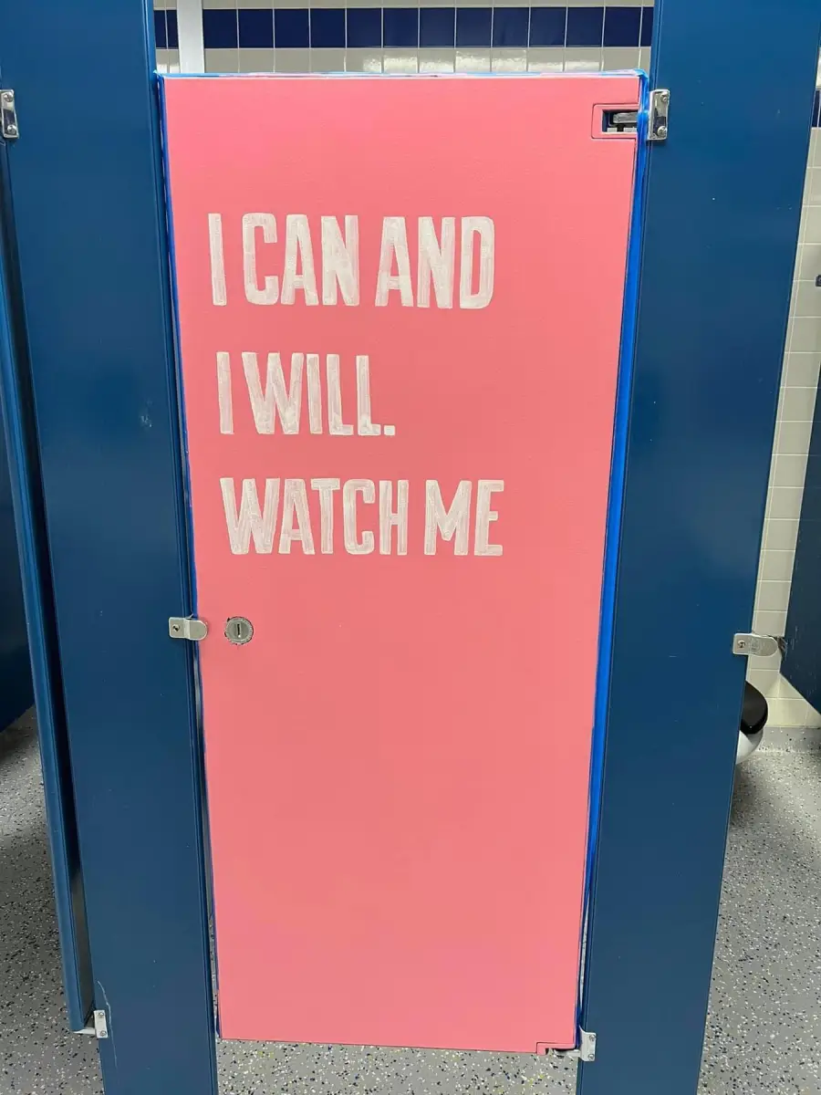 A pink bathroom stall door with the words “I CAN AND I WILL. WATCH ME” painted in bold white letters. The surrounding stall dividers are blue.