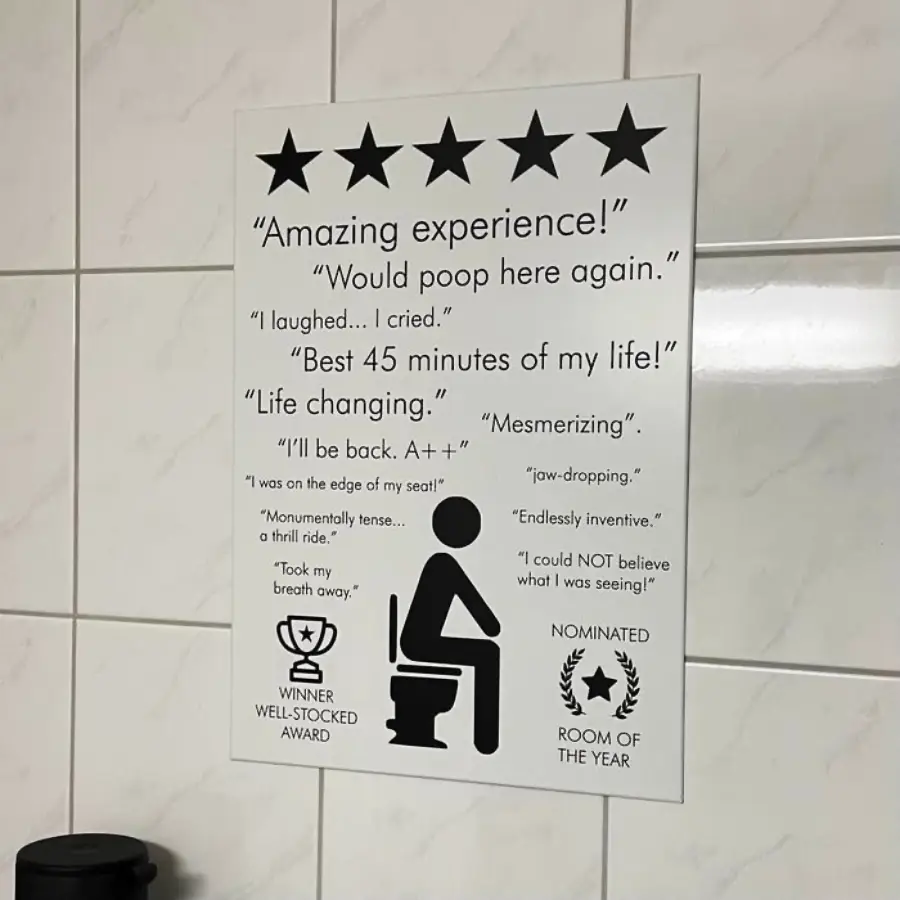 White bathroom wall with a humorous poster featuring a person on a toilet, five stars, and funny fake reviews like “Would poop here again” and “Best 45 minutes of my life!” along with playful award icons.