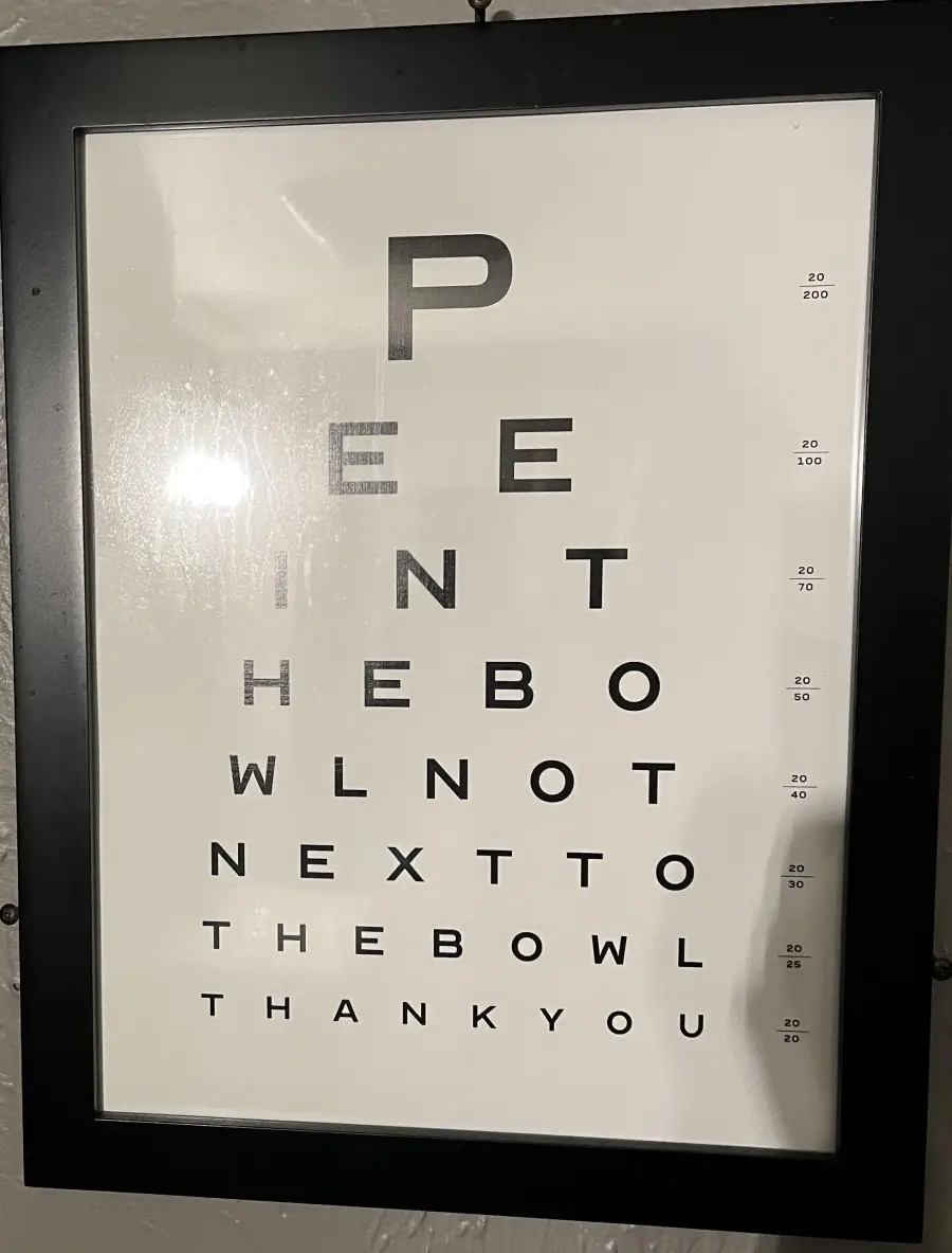 A framed chart resembling an eye test reads, “Pee in the bowl not next to the bowl thank you,” with letters decreasing in size from top to bottom.