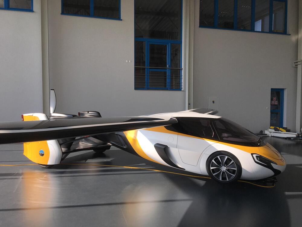 A sleek prototype flying car with yellow and white accents is parked indoors, showcasing extended wings and a streamlined design, blending features of both a sports car and a small aircraft.