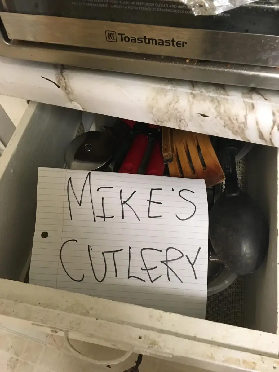 A kitchen drawer with utensils and a handwritten sign on lined paper that reads “MIKE’S CUTLERY.” The area above the drawer appears dirty and stained.