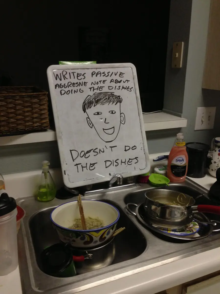 A whiteboard over a kitchen sink reads: “WRITES PASSIVE AGGRESSIVE NOTE ABOUT DOING THE DISHES. DOESN’T DO THE DISHES,” with a doodle of a face. Dirty dishes are piled in the sink below.