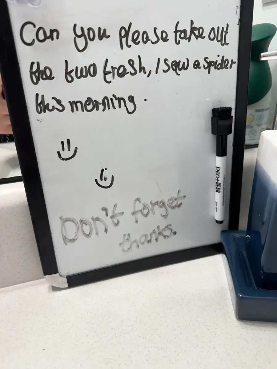 A whiteboard with a handwritten note: “Can you please take out the two trash, I saw a spider this morning,” plus smiley faces and “Don’t forget thanks” written below. A marker and soap dispenser are nearby.