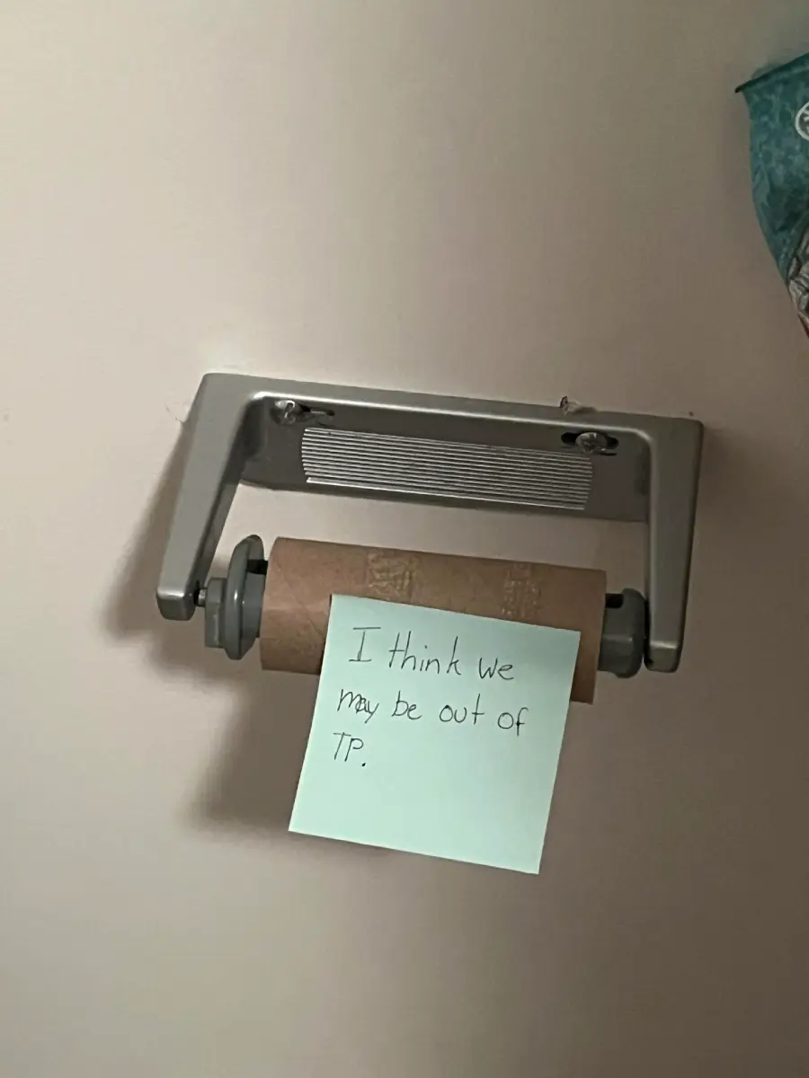 An empty toilet paper roll with a sticky note that says, “I think we may be out of TP,” attached to the holder on a beige wall.