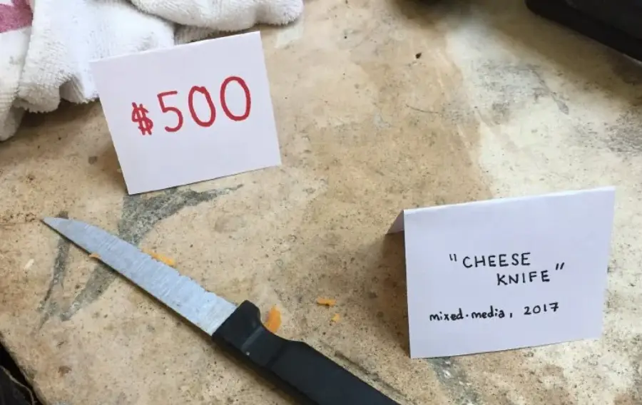 A kitchen knife with cheese on the blade lies on a counter. A card reads ‘“CHEESE KNIFE” mixed media, 2017.’ Another card shows a price of $500 in red.