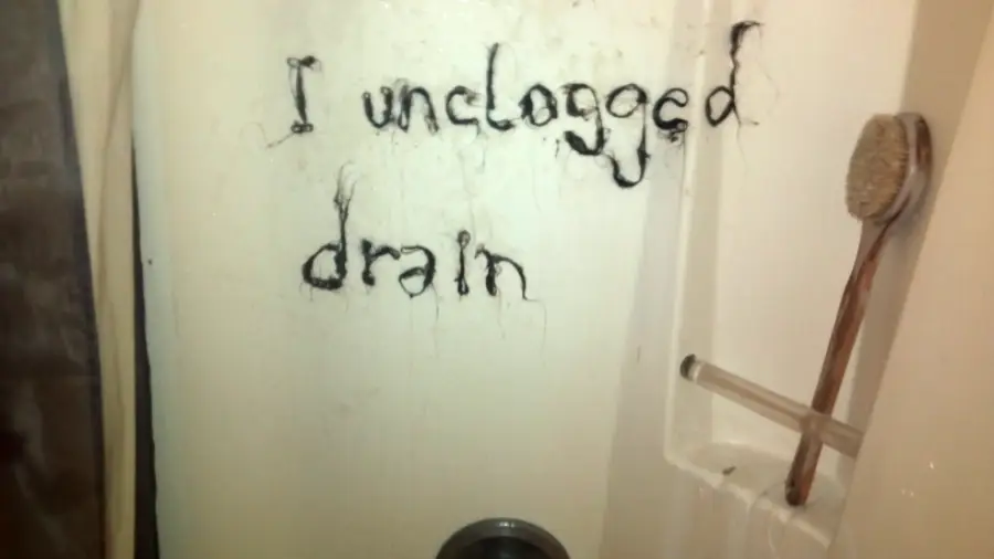 Hair spelling out “I unclogged drain” is stuck to a shower wall above the drain. A brush and a razor are in the shower caddy on the right.