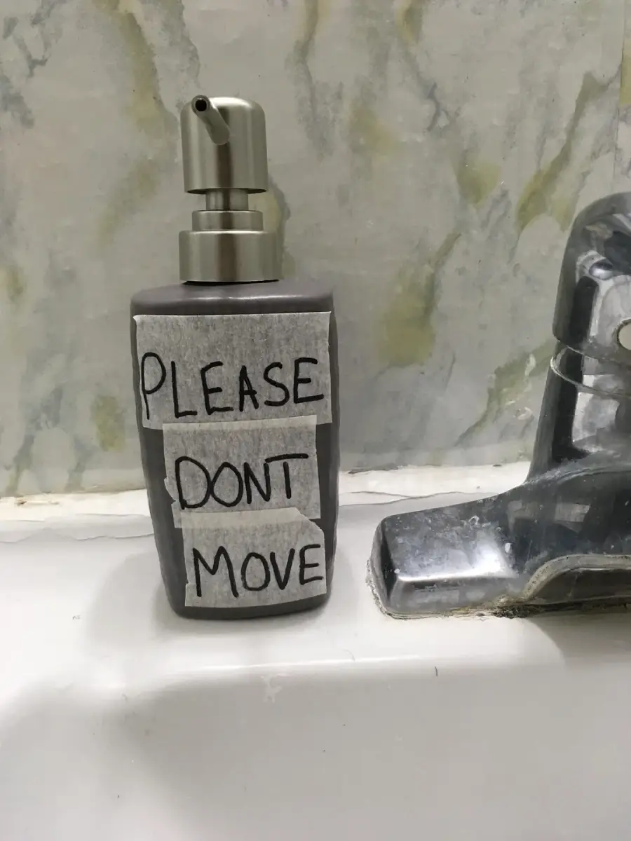 A soap dispenser labeled with taped-on paper reading “PLEASE DON’T MOVE” sits next to a faucet on a bathroom sink with a marbled wall in the background.
