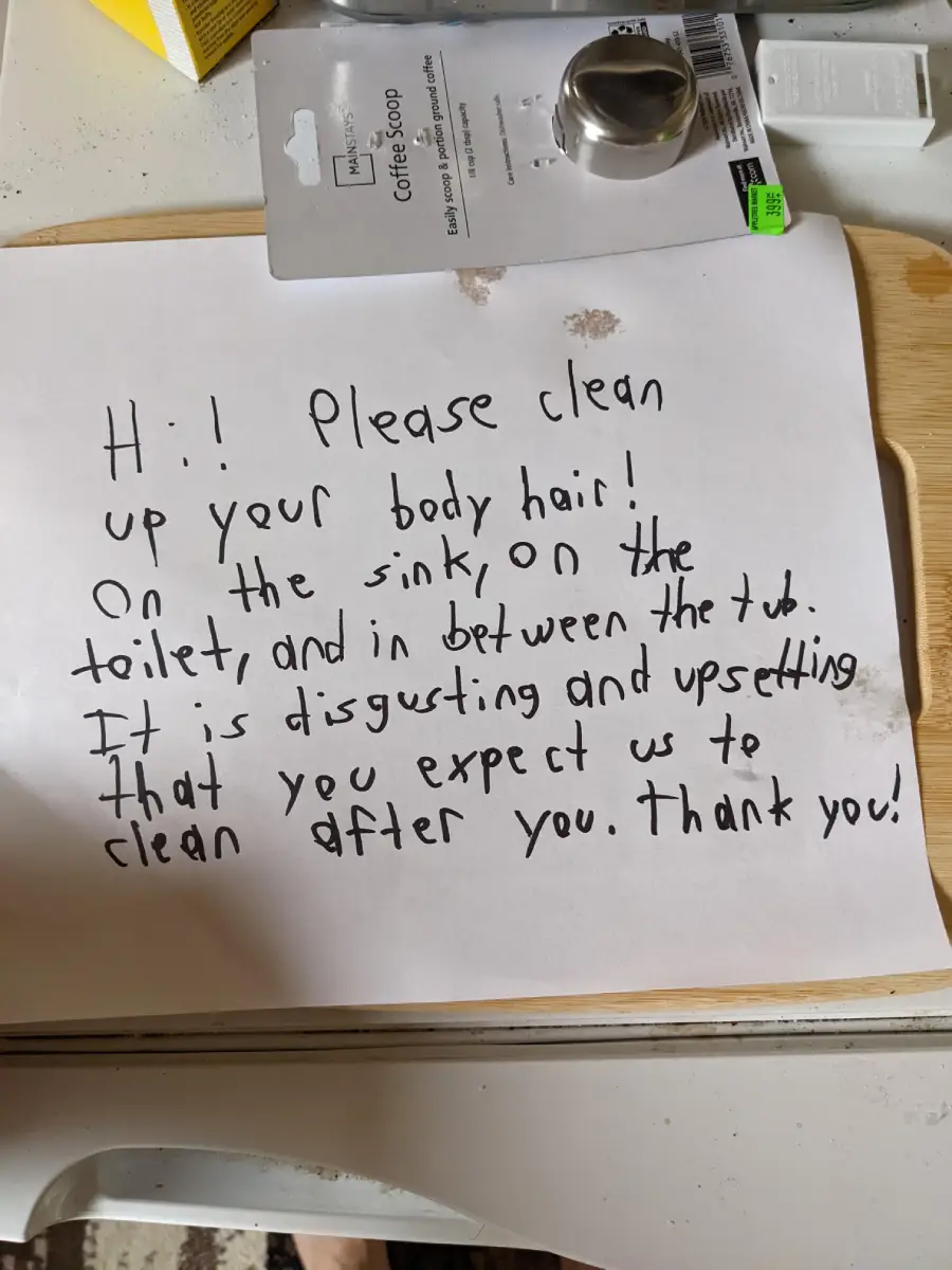 A handwritten note on white paper asks others to clean up body hair from the sink, toilet, and tub, calling it disgusting and upsetting. The note ends with “Thank you!” The paper rests on a table with various items nearby.