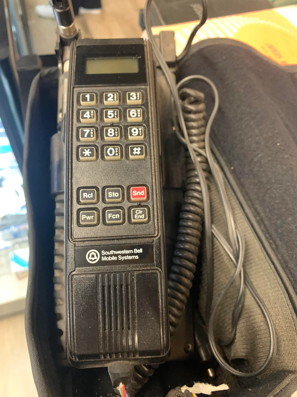 A vintage Southwestern Bell Mobile Systems car phone with a keypad, digital display, and coiled cord resting in a carrying case.