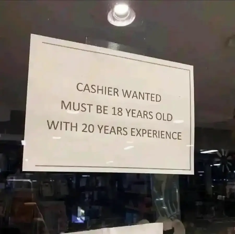 A sign reads: “CASHIER WANTED. MUST BE 18 YEARS OLD WITH 20 YEARS EXPERIENCE” and is posted on a glass door or window.