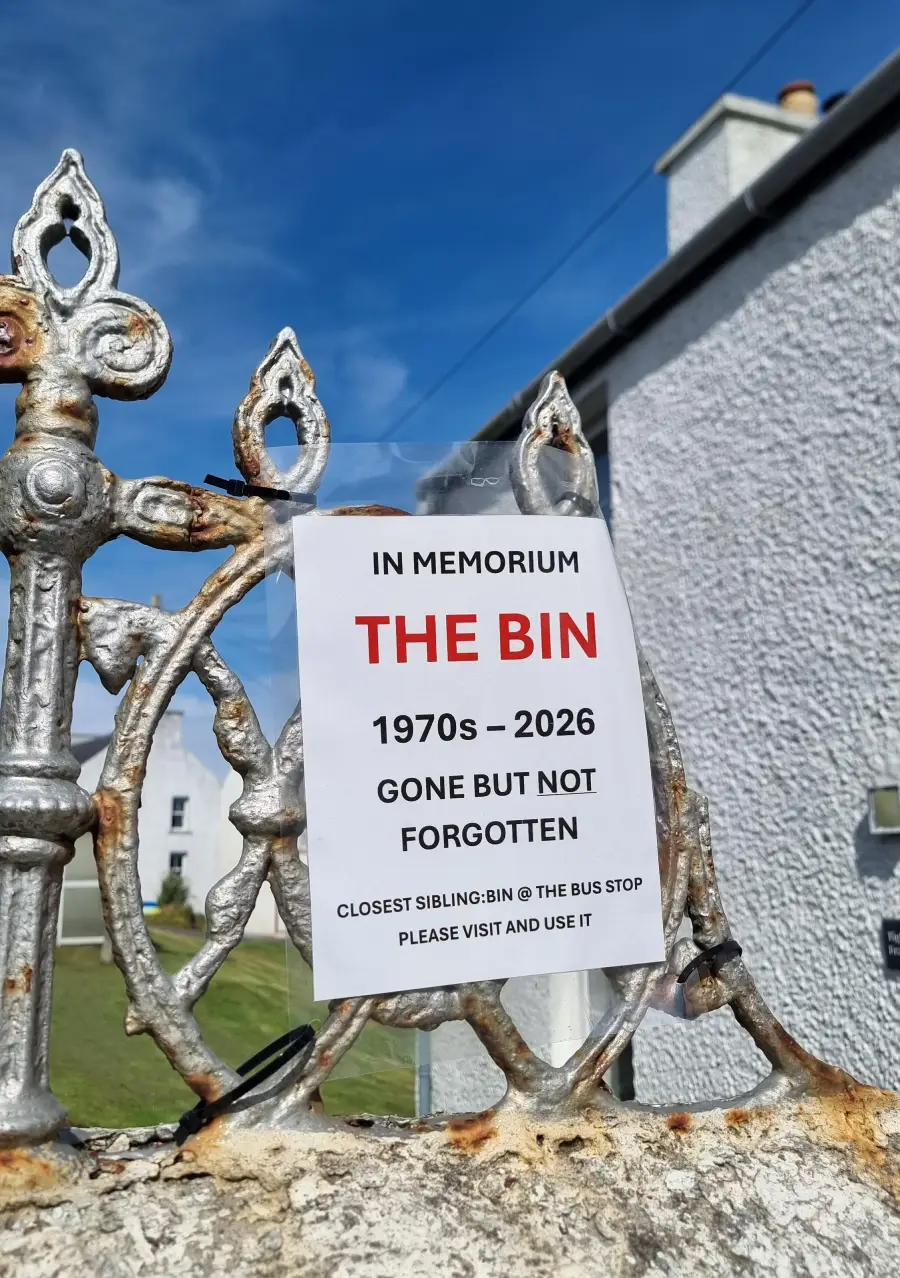 A printed sign on a decorative metal fence reads, “IN MEMORIUM THE BIN 1970s – 2026 GONE BUT NOT FORGOTTEN,” with instructions to find the closest bin at the bus stop.