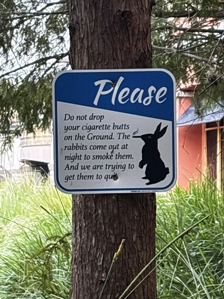 A sign on a tree says, “Please do not drop your cigarette butts on the ground. The rabbits come out at night to smoke them. And we are trying to get them to quit.” A black rabbit silhouette is shown.