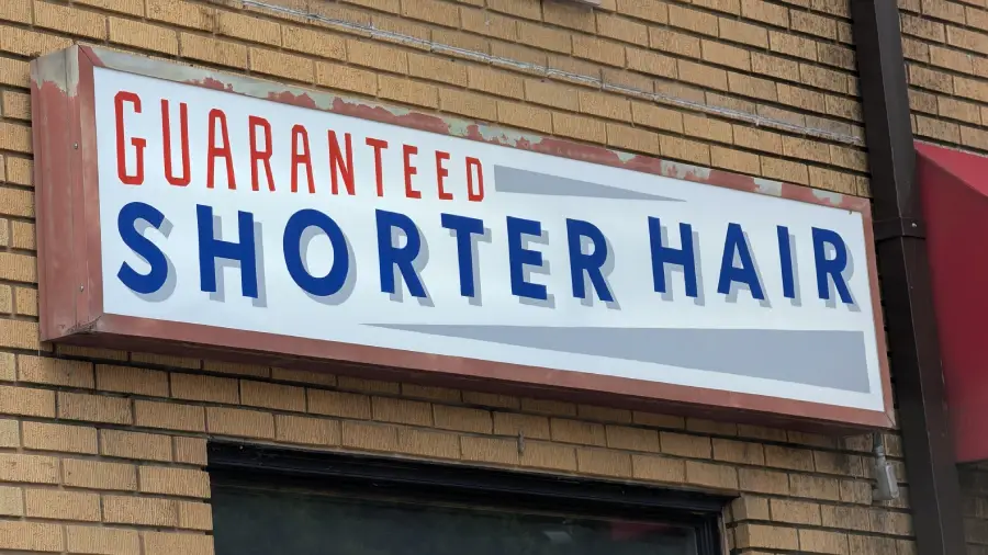 A white sign on a brick wall reads “Guaranteed Shorter Hair” in blue and red letters outside what appears to be a barbershop or hair salon.