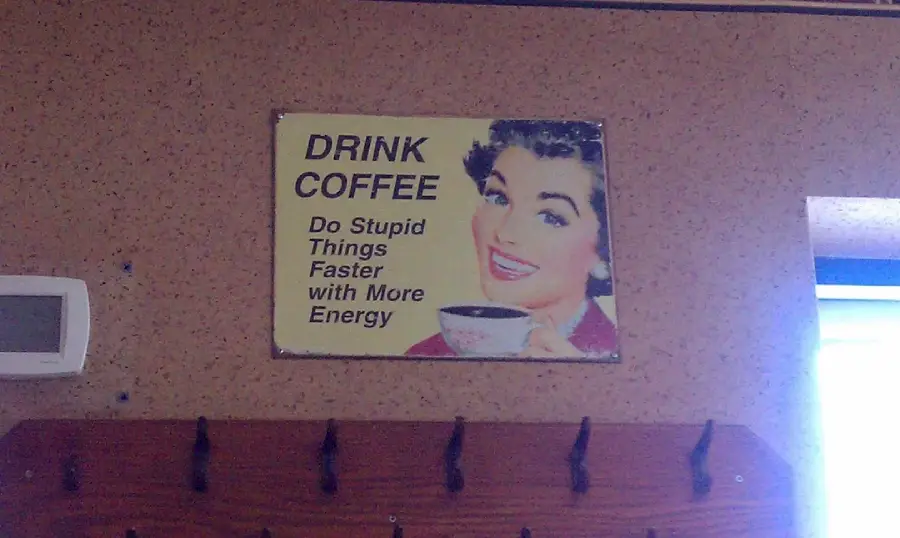 A vintage-style sign with a smiling woman holding a coffee cup. The sign reads: “DRINK COFFEE. Do Stupid Things Faster with More Energy.” The background is a light brown wall with coat hooks below the sign.