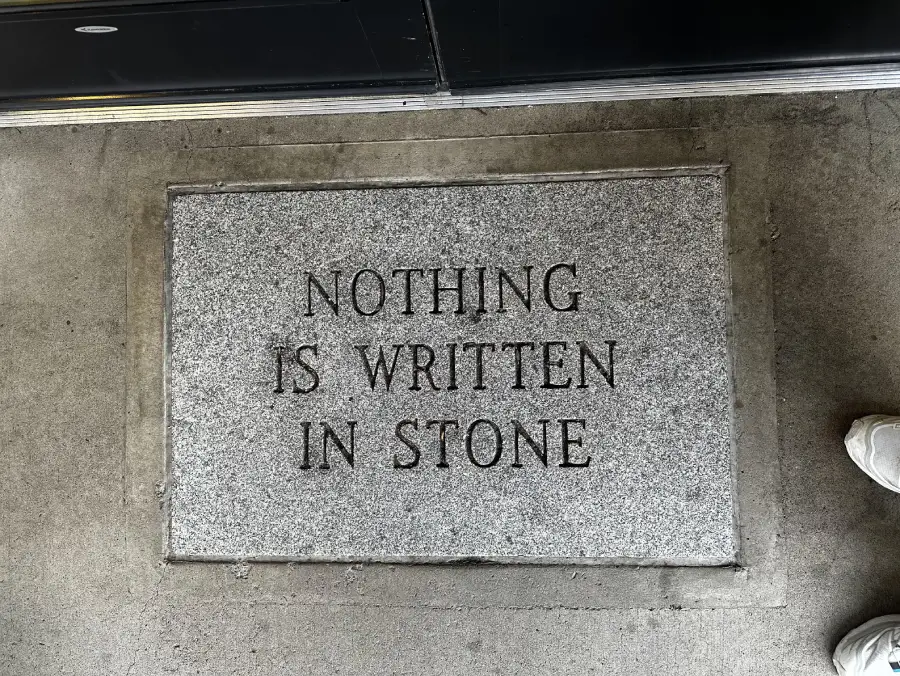 A stone slab on the ground has the phrase “NOTHING IS WRITTEN IN STONE” engraved on it. Part of a white shoe is visible in the lower right corner.