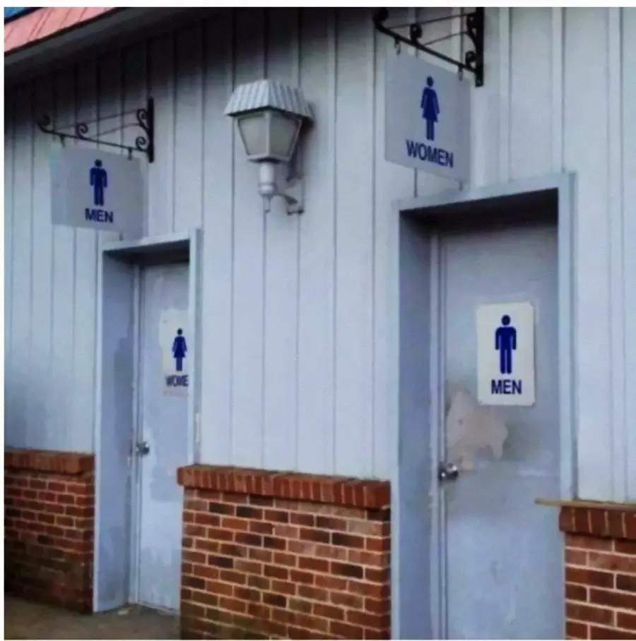 Two restroom doors on a brick building; the door labeled “MEN” has a sign above it reading “WOMEN,” and the door labeled “WOMEN” has a sign above it reading “MEN.”
