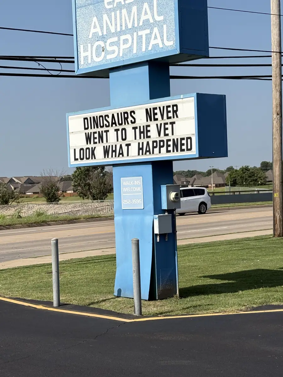A veterinary clinic sign reads, “DINOSAURS NEVER WENT TO THE VET. LOOK WHAT HAPPENED,” with houses, a street, and a white SUV in the background.