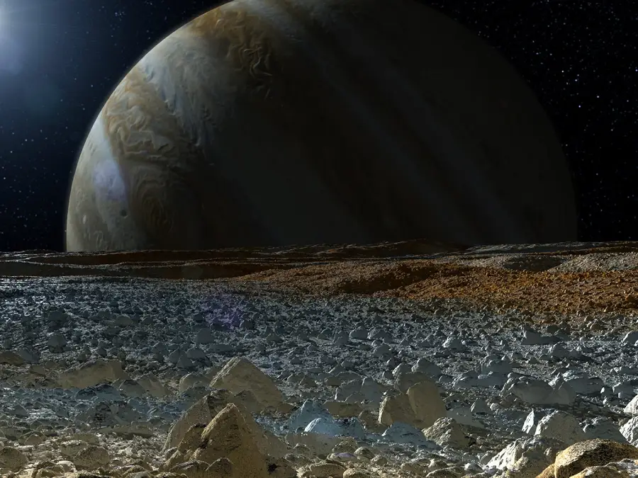 Rocky, icy landscape with large boulders under a dark sky. A giant planet with visible bands dominates the horizon, with a bright light source shining from the left.