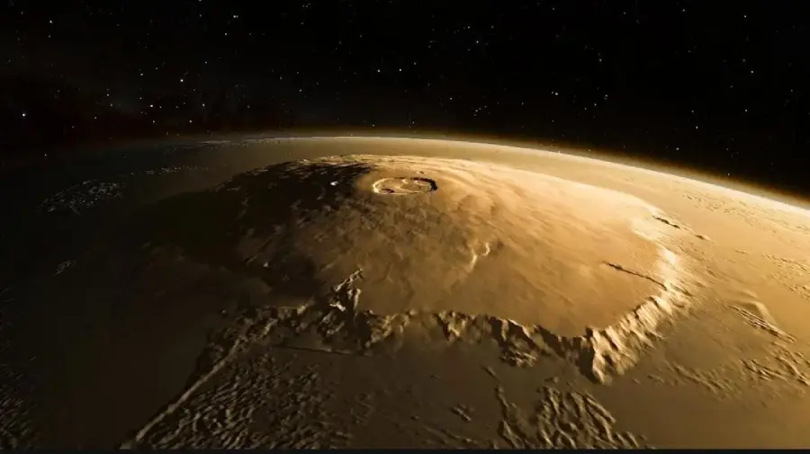 A wide view of Olympus Mons, a massive shield volcano on Mars, showing its large, circular caldera and gently sloping sides under dim sunlight with space in the background.