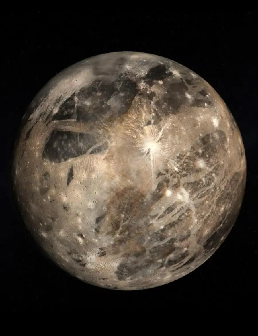 A detailed image of Ganymede, Jupiter’s largest moon, showing its heavily cratered, icy surface with dark and light regions, streaks, and a prominent bright impact crater near the center, set against the blackness of space.