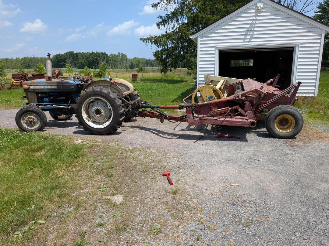 A blue tractor is hitched to a large, red piece of farm equipment on a gravel driveway in front of a white shed, with grassy fields and trees in the background. A red tool lies on the ground nearby.