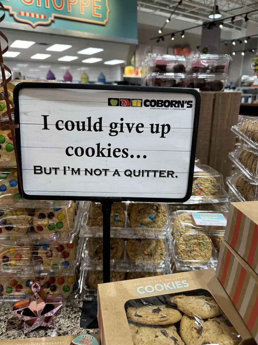 A store display shows a sign reading, “I could give up cookies... but I’m not a quitter,” surrounded by packages of cookies and baked goods.