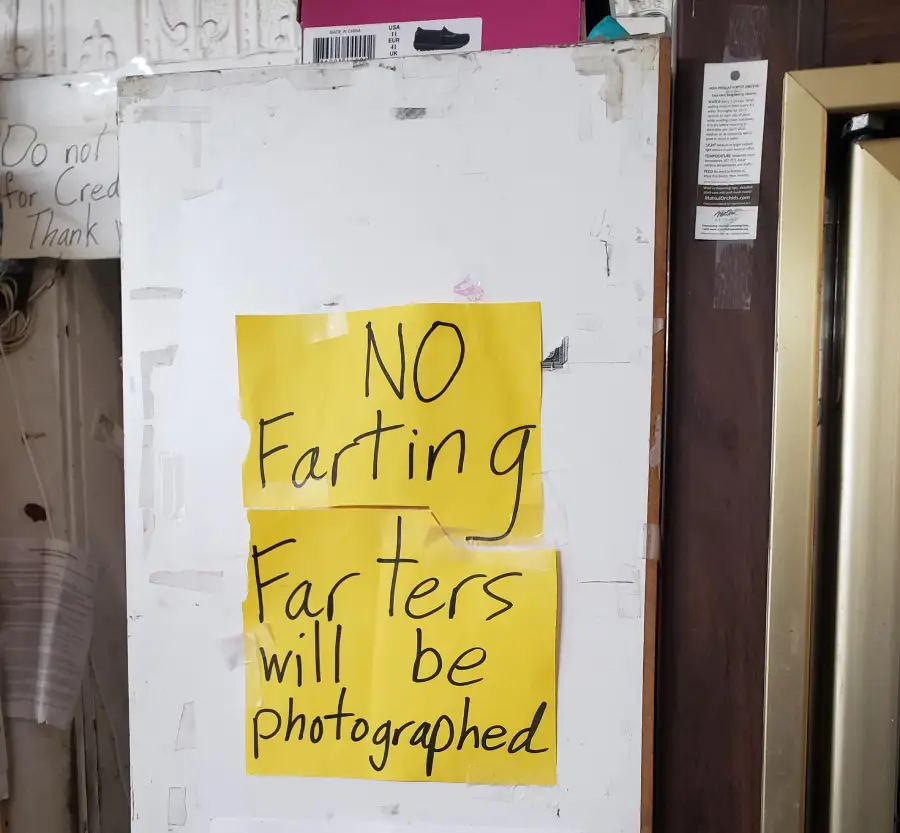 A whiteboard with a yellow sign that says, “NO Farting Farters will be photographed,” written in black marker, taped in a cluttered indoor space.
