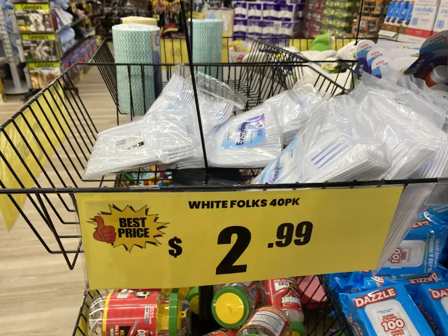 Plastic forks in clear packaging are displayed in a wire basket above a yellow sign that reads “WHITE FOLKS 40PK” with a price of $2.99 at a store. Other household items are visible in the background.